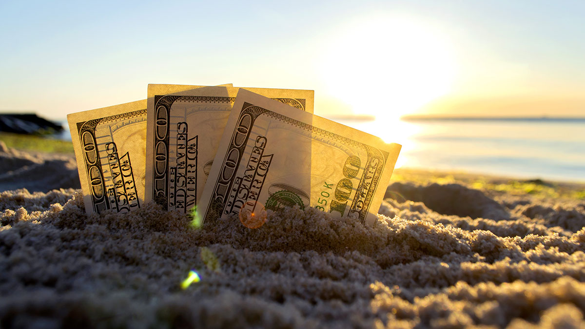 Dollar bills half buried in the sand backdrop of a beach sunset