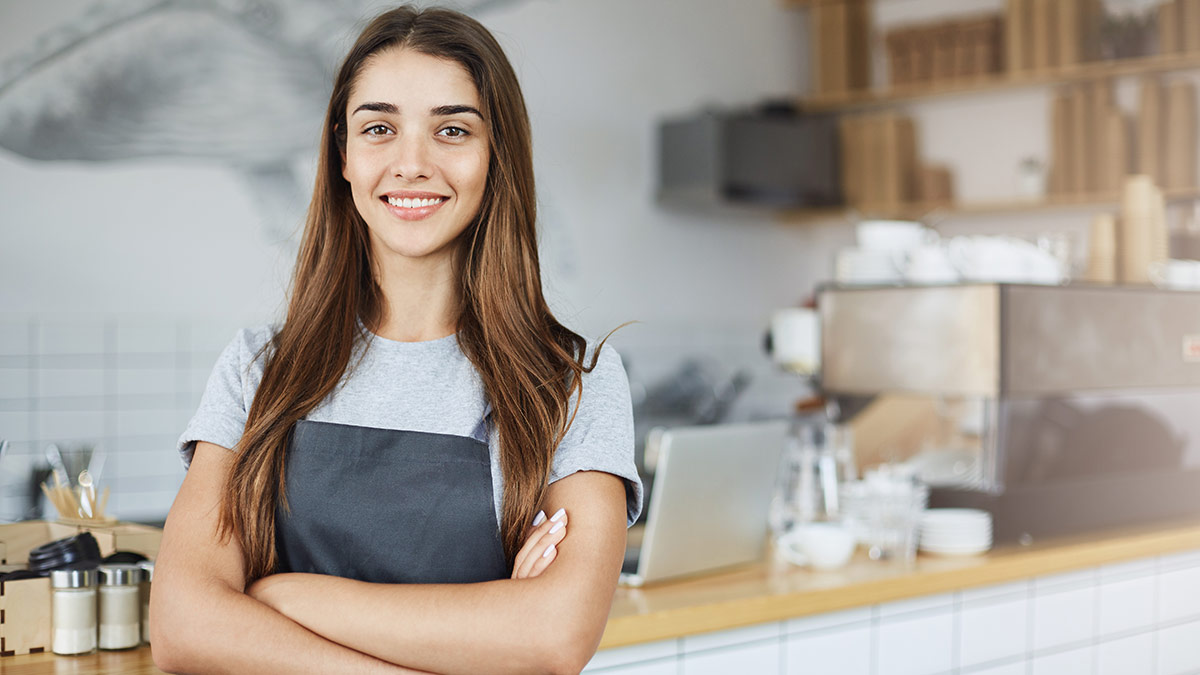 teenage girl with barista apron arms folded with coffee shop in background