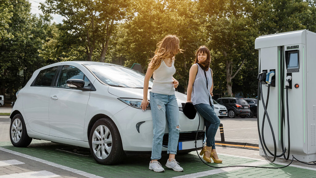electric vehicle being charged at a station with two young women talking leaning on car while they wait for it to charge