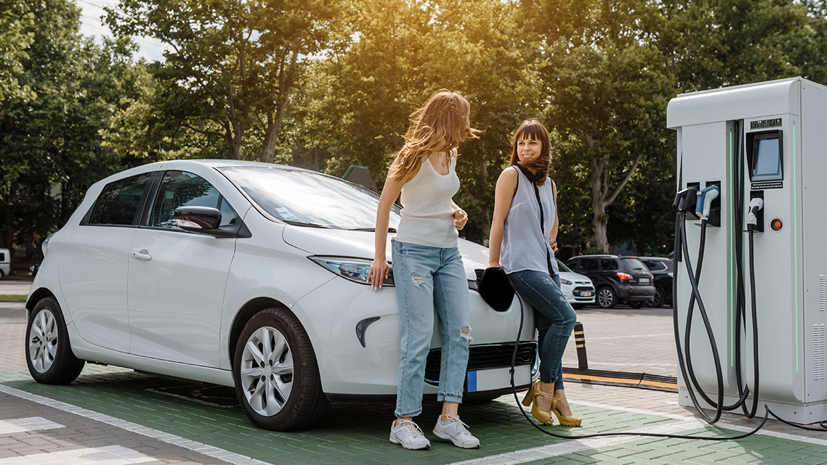 electric vehicle being charged at a station with two young women talking leaning on car while they wait for it to charge