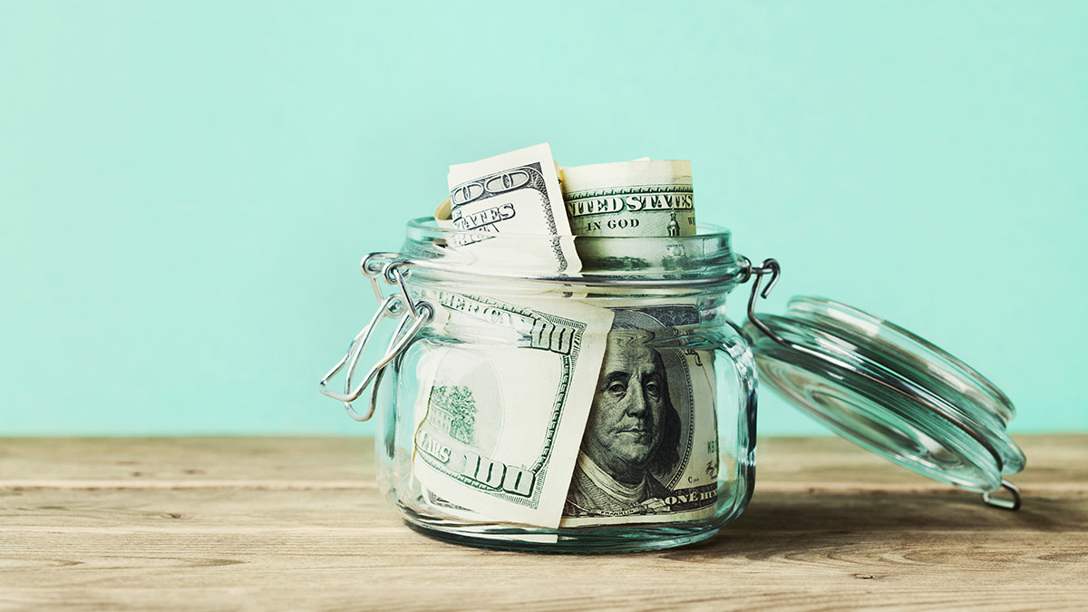 money in a tips jar on a wooden table with a turquoise blue background